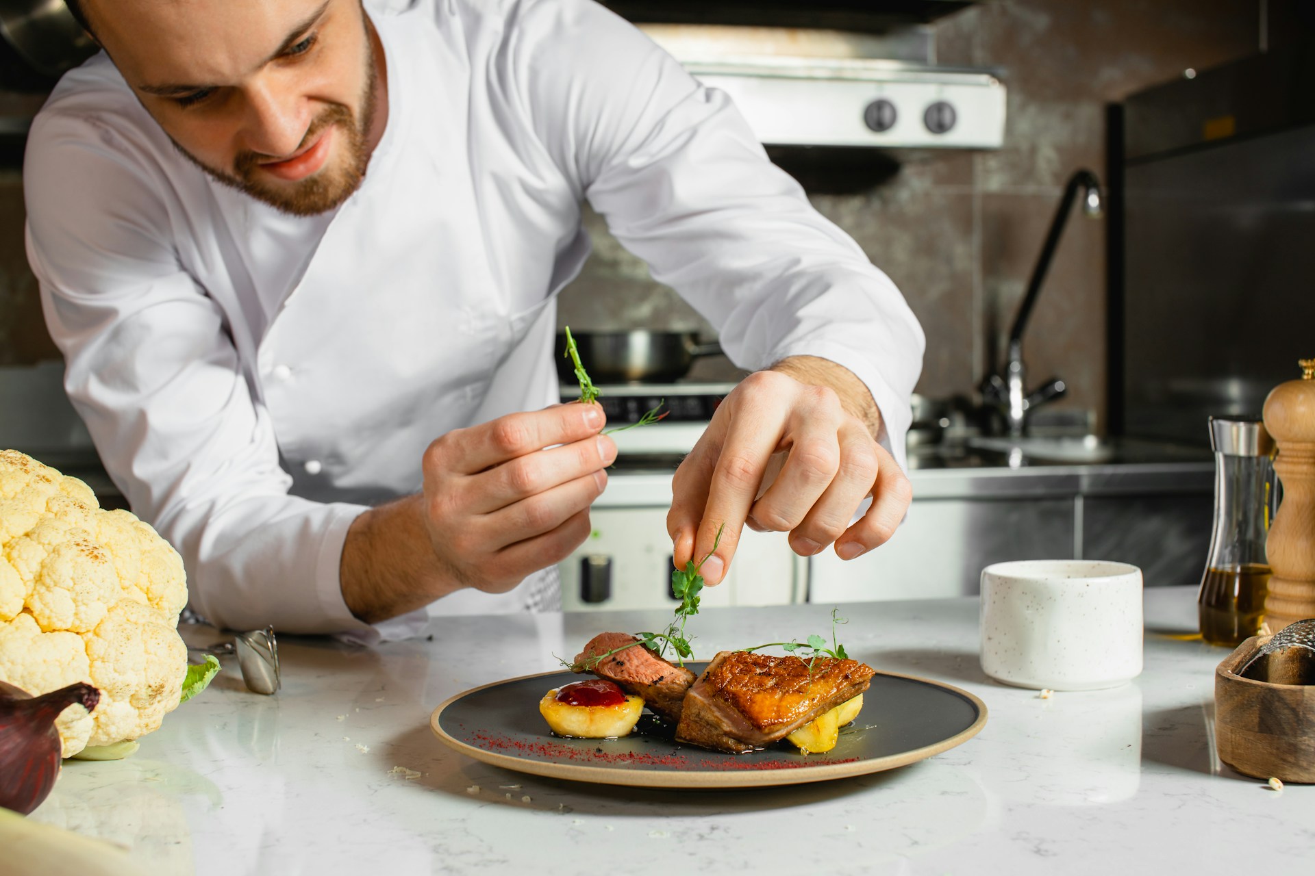 Chef garnishing a plated dish featuring meat and vegetables in a modern kitchen setting.