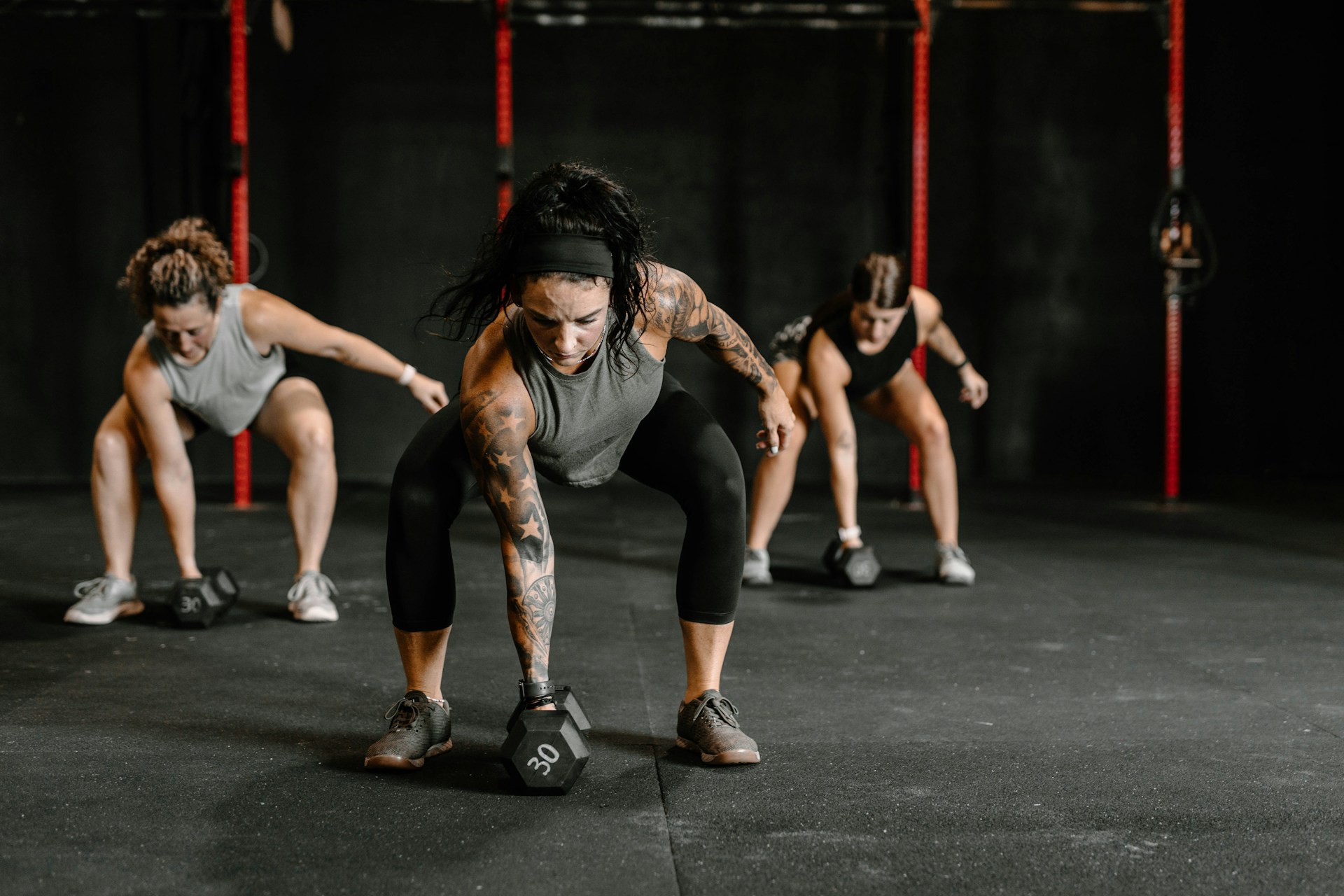 Three women perform strength exercises with dumbbells in a gym setting.