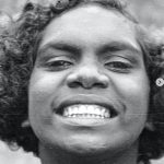 Young person with curly hair smiling broadly, showcasing bright white teeth against a plain background.