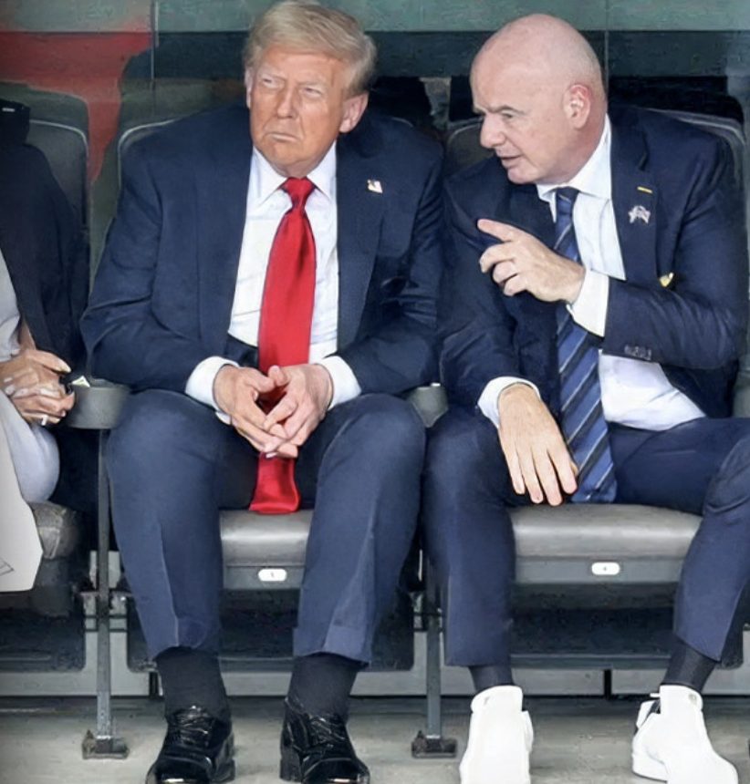Donald Trump and a man in a suit converse while seated in a stadium.
