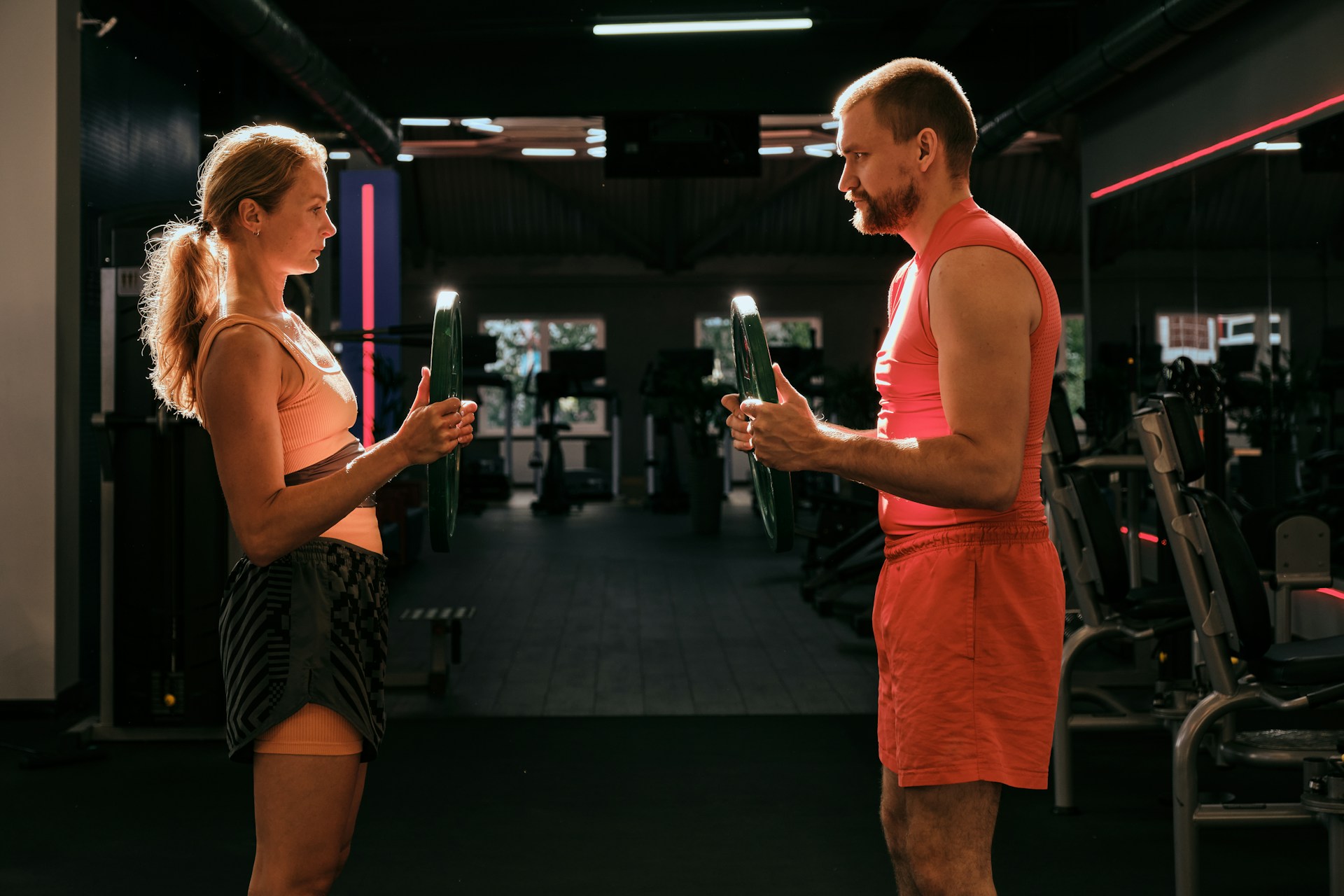 Two people in athletic attire hold weight plates while facing each other in a gym setting.