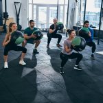 Five individuals perform squats with medicine balls in a modern gym, showcasing teamwork and fitness.
