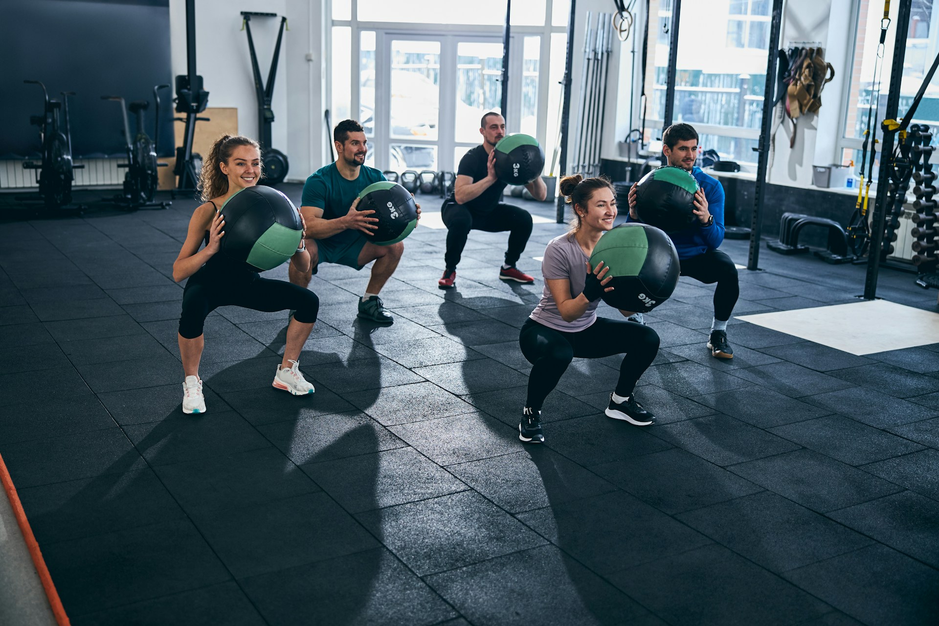 Five individuals perform squats with medicine balls in a modern gym, showcasing teamwork and fitness.