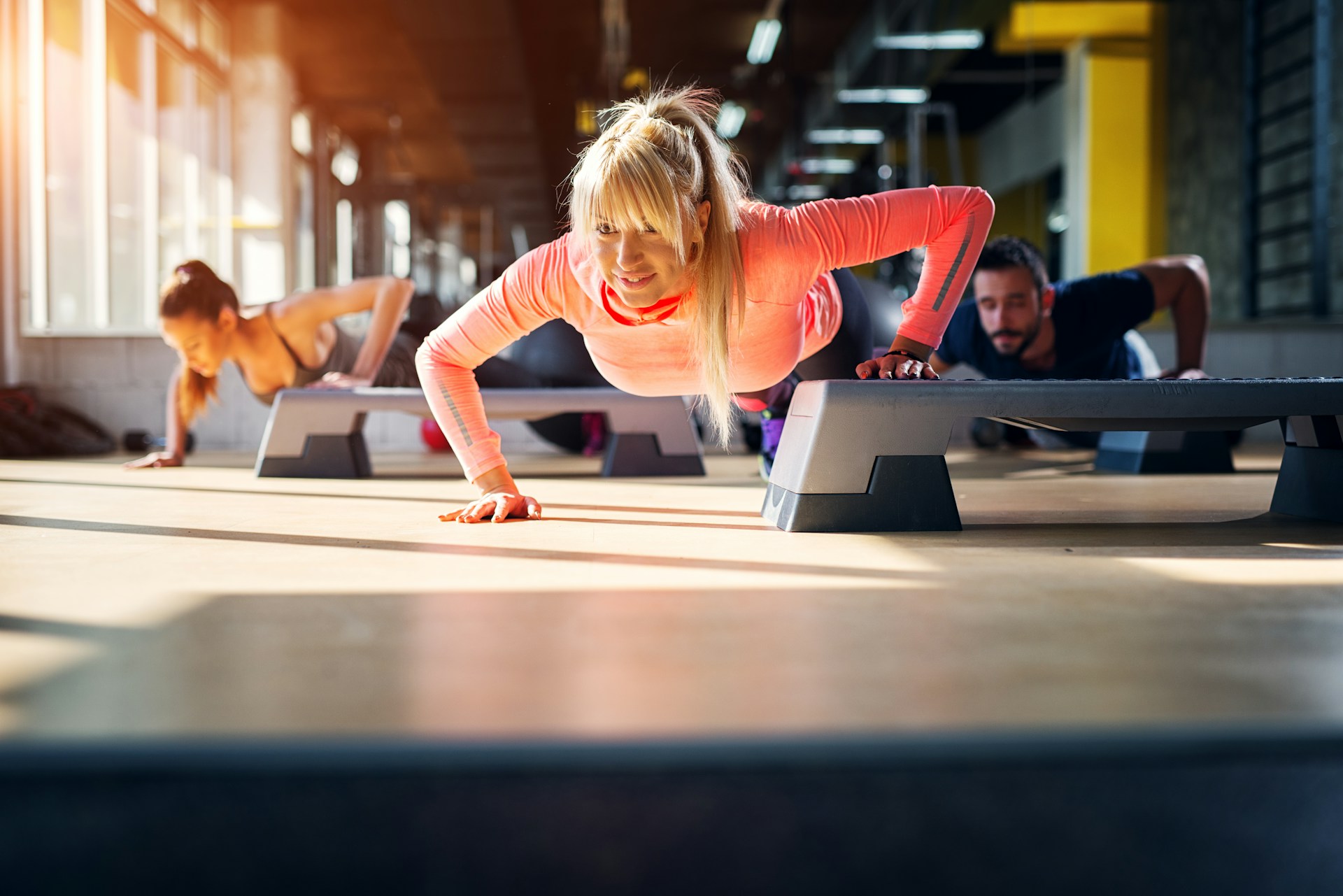 Three individuals perform push-ups on fitness steps in a brightly lit gym.