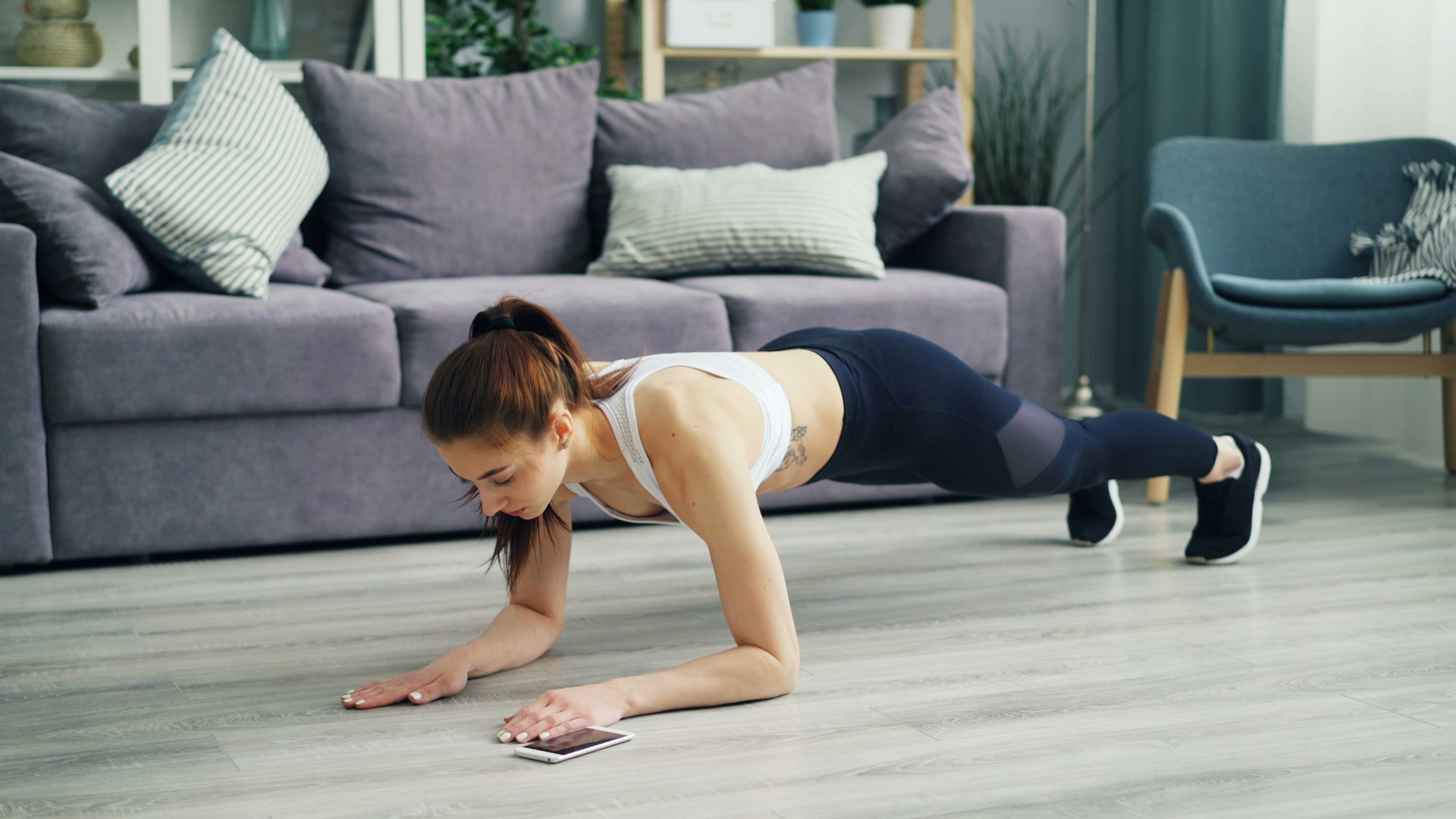 Woman in a plank position on a floor, exercising in a cozy living room with a sofa and plants.